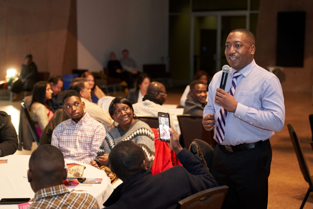 Man speaking into a microphone while individuals watch seated at a table.